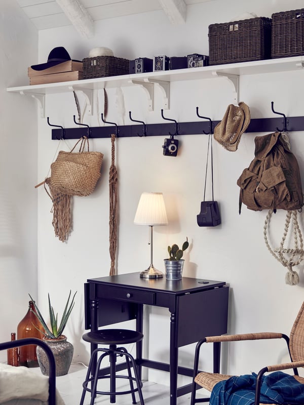 On a beige wall hangs a white and black traditional-style hat rack with jackets, a bag, an umbrella, a basket and hats.