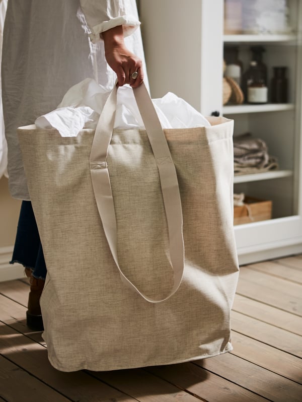 Una persona vicino a una vetrina porta un sacco per il bucato PURRPINGLA beige tenendolo per i manici corti - IKEA.