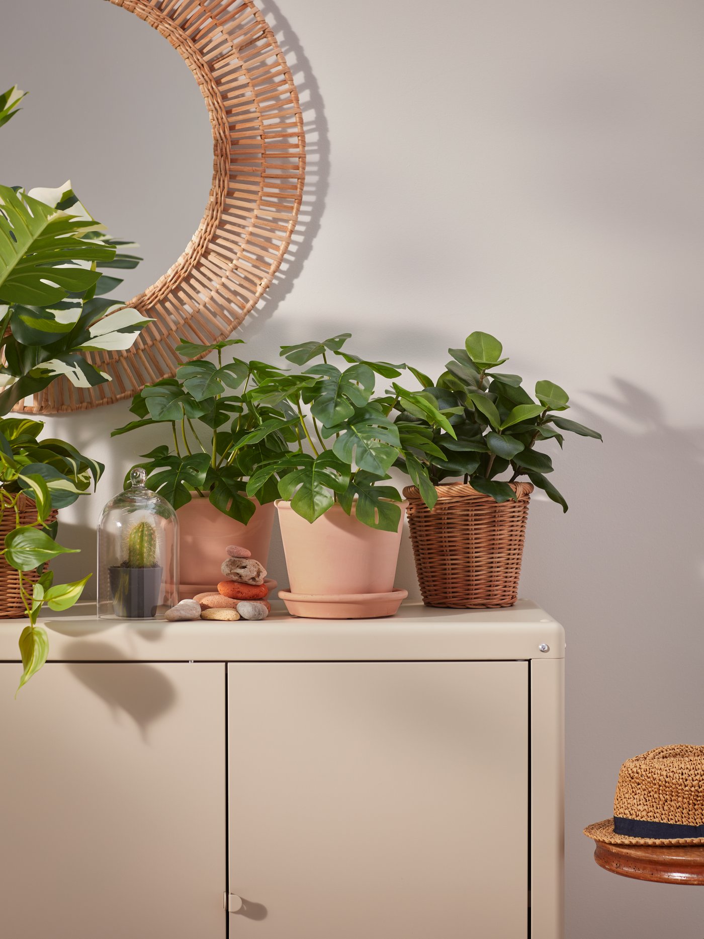 Plantas artificiales en macetas sobre un armario beige. Encima del armario hay un espejo de pared redondo con borde decorativo.
