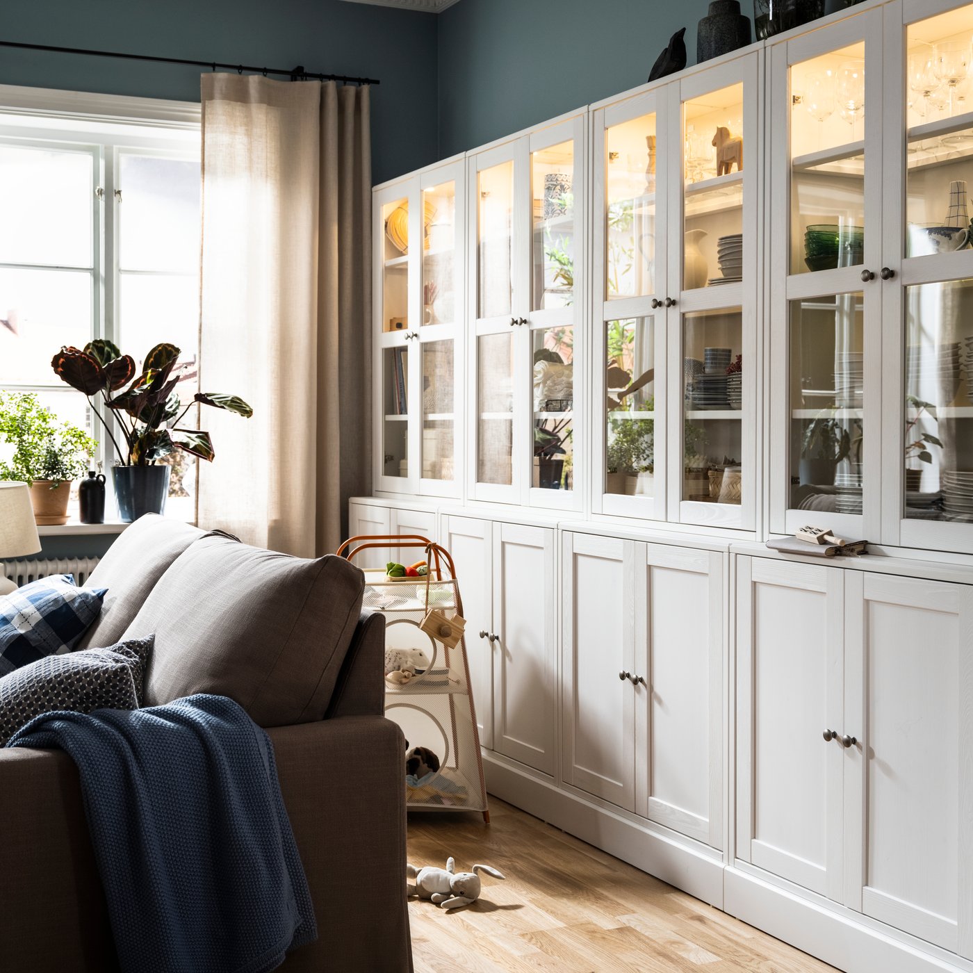 Some white HAVSTA storage combinations with glass-doors stand against a wall behind a grey-brown HYLTARP sofa-bed.