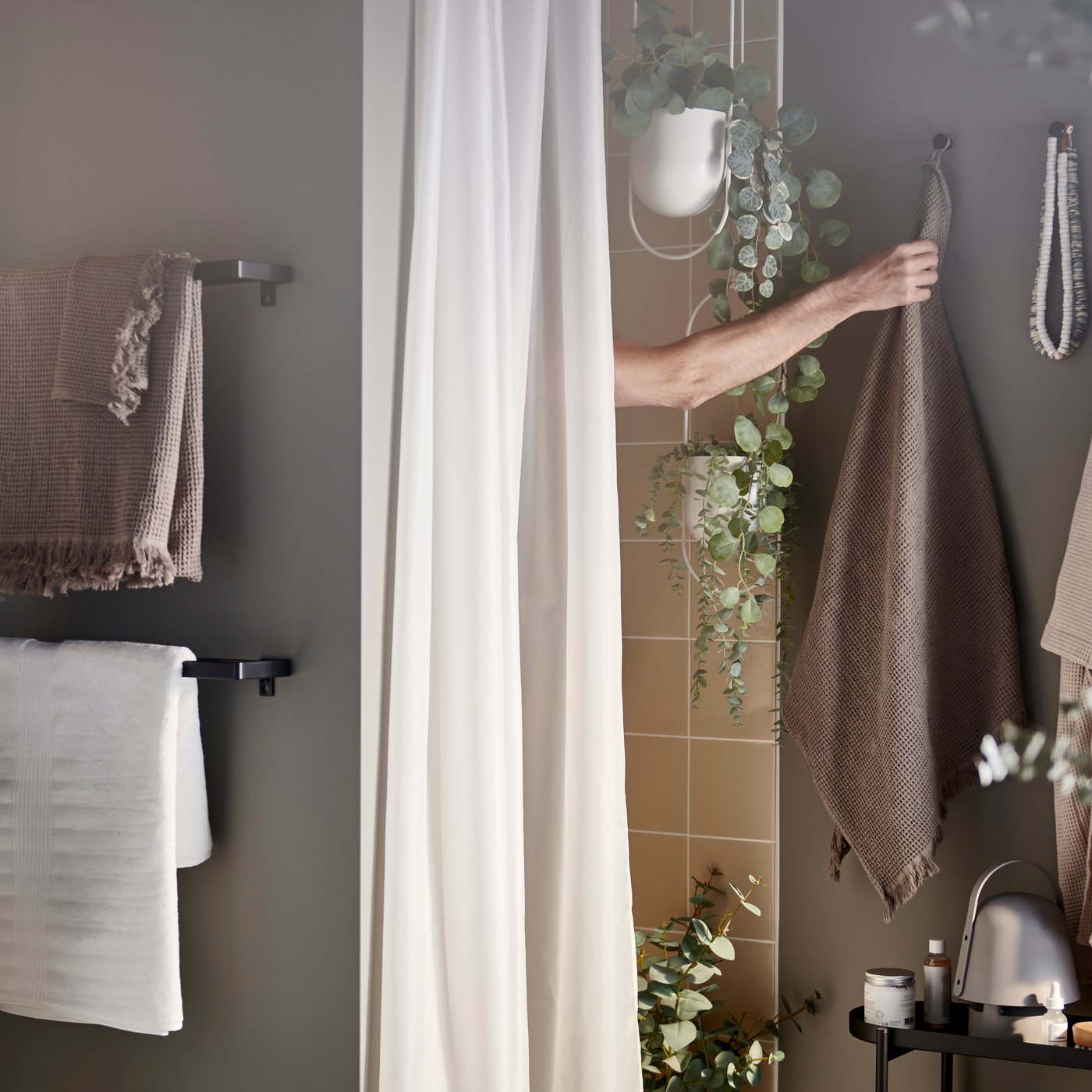 Een arm reikt vanuit de douche naar een handdoek in lichtgrijs/bruin die aan een haak hangt. We zien ook een douchegordijn en kunstplanten in potten.