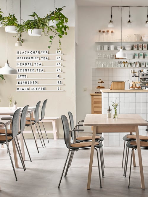 Salle à manger avec plusieurs tables rectangulaires en bouleau massif, plusieurs chaises avec accoudoirs et un banc beige.