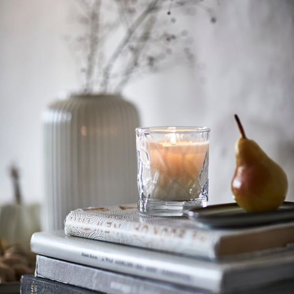 A scented HÖSTAGILLE candle in a glass jar is placed on stacked books beside a decorative vase and a pear.