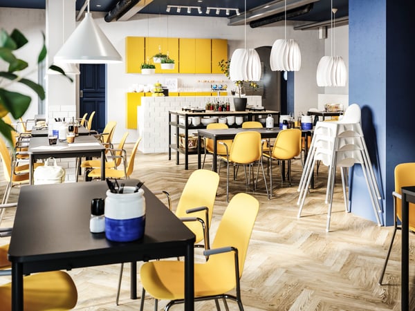 A bright restaurant with dark yellow chairs, black tables, white junior chairs, yellow cabinets and white pendant lamps.