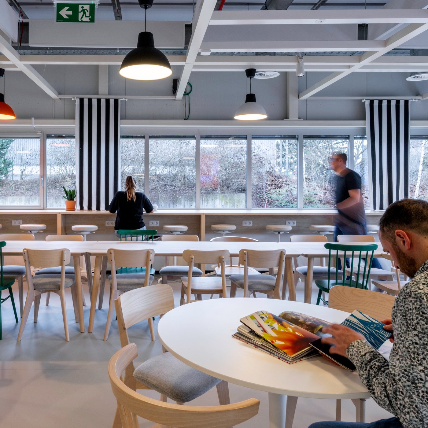 Spacieuse zone de restauration de bureau avec du mobilier en bois clair, des chaises d’accent vertes et des suspensions colorées, où des collaborateurs font une pause.