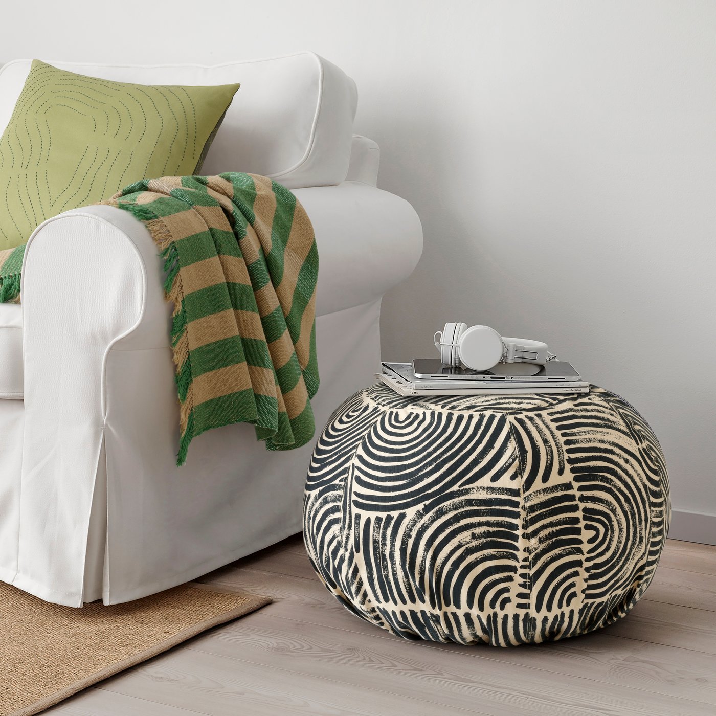 A FRÖDD black/patterned floor cushion next to a white sofa. A FRÖDD green striped throw hangs off the arm of the white sofa.