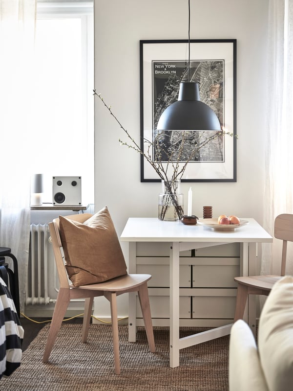 En flexibel matplats med ett vitt slagbord och två stolar i ljust trä. Ovanför matbordet hänger en svart lampa och bakom syns en stor tavla med stadsobjekt.