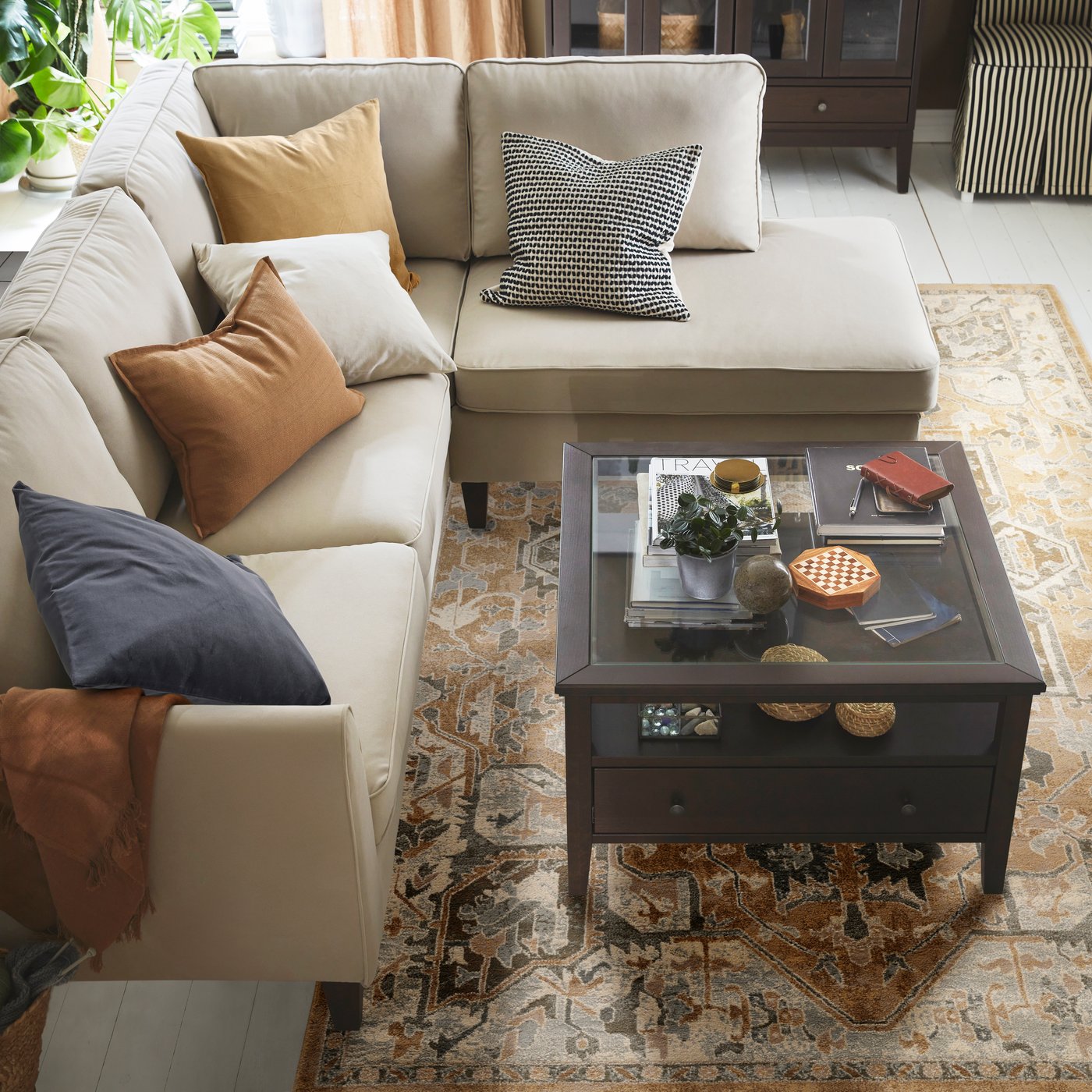 Ein beiger JÄRNVÄG Teppich mit orientalischem Muster liegt unter einem beigen Ecksofa. Vor dem Sofa steht ein schwarzbrauner IDANÄS Couchtisch. Auf dem Sofa befinden sich SANELA Kissen in verschiedenen Farben, wie dunkelgrau und goldgelb.