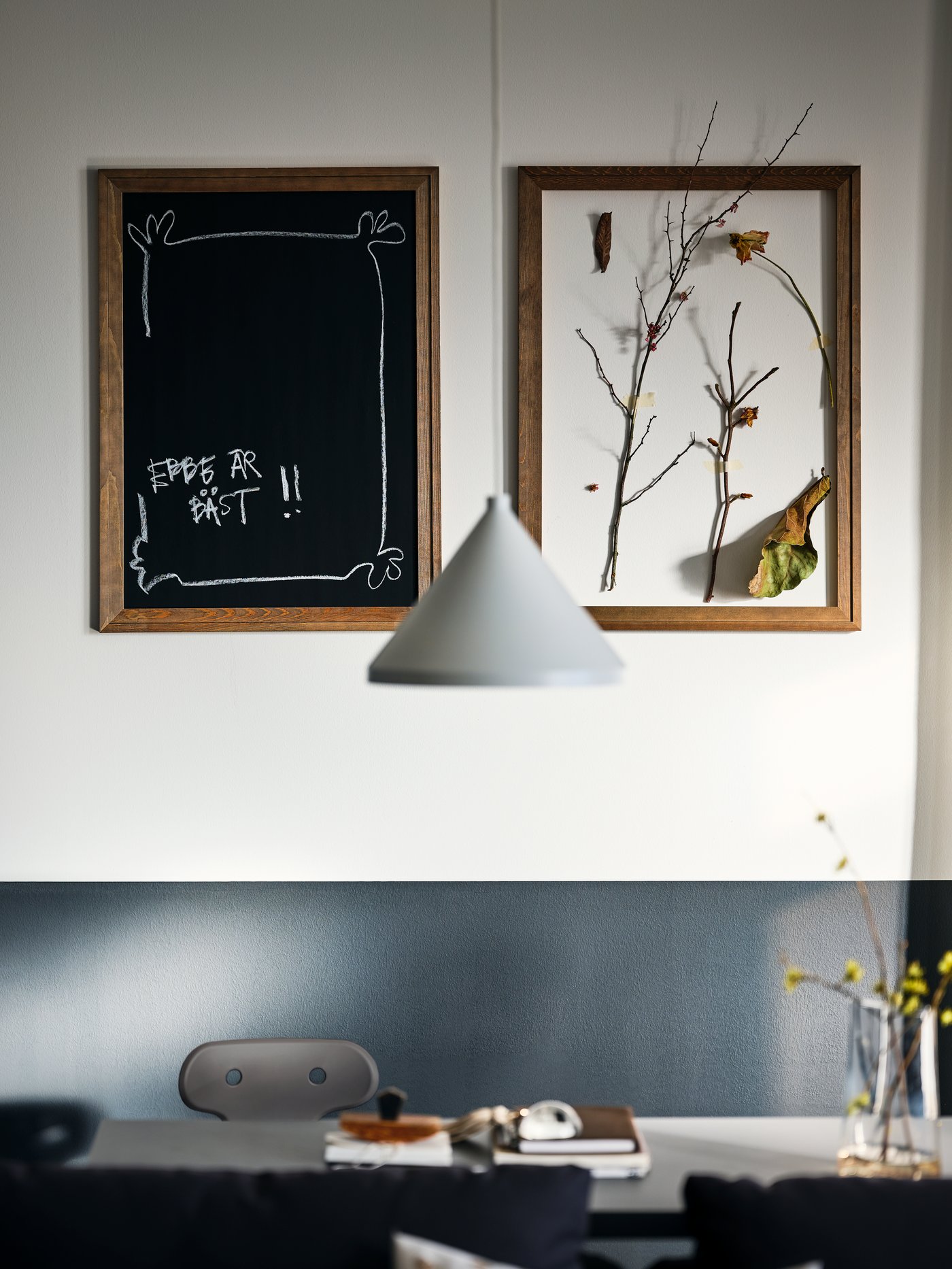 A dining area with a white NÄVLINGE pendant above it, a MOLTE desk chair, and frames with a blackboard and art on the wall.