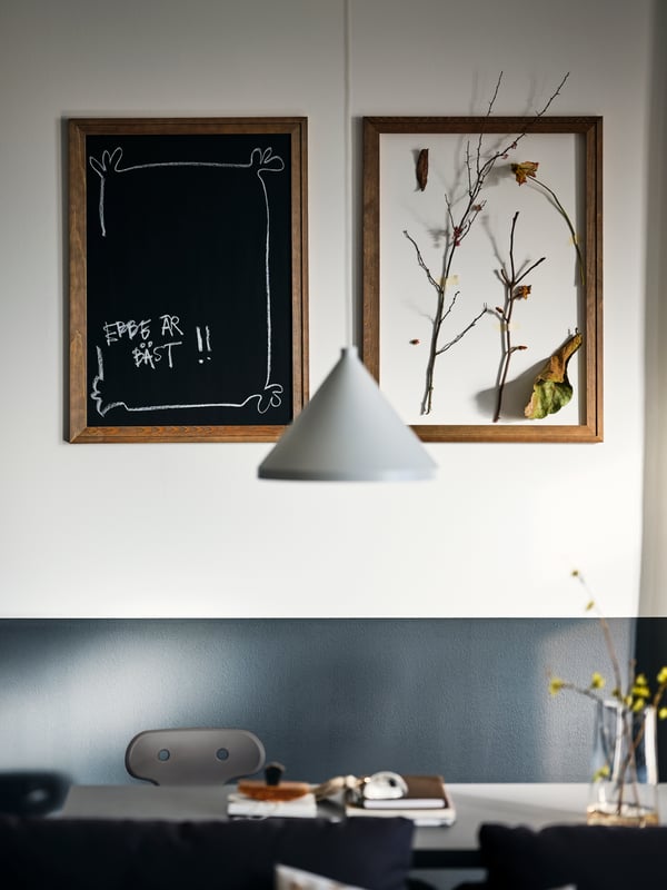 A dining area with a white NÄVLINGE pendant above it, a MOLTE desk chair, and frames with a blackboard and art on the wall.