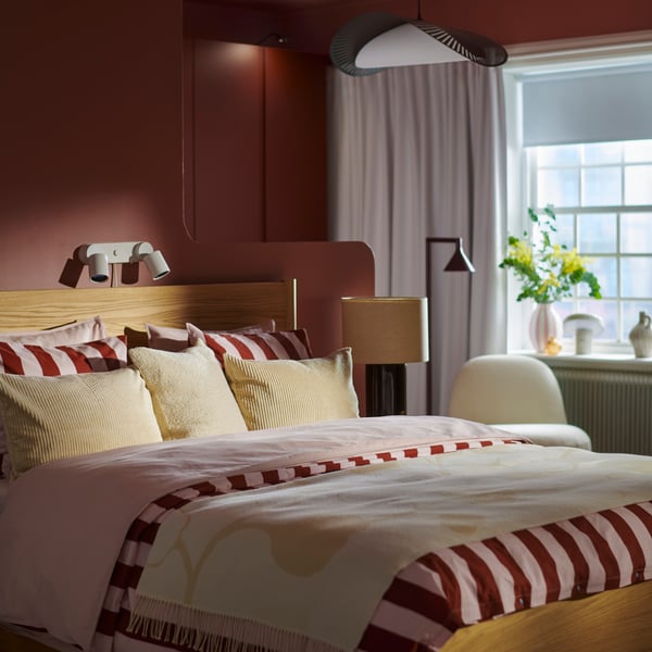 A bedroom with oak veneer TONSTAD bed made with striped SLÅNHÖSTMAL bedding and cream pillows and throw with a window behind.