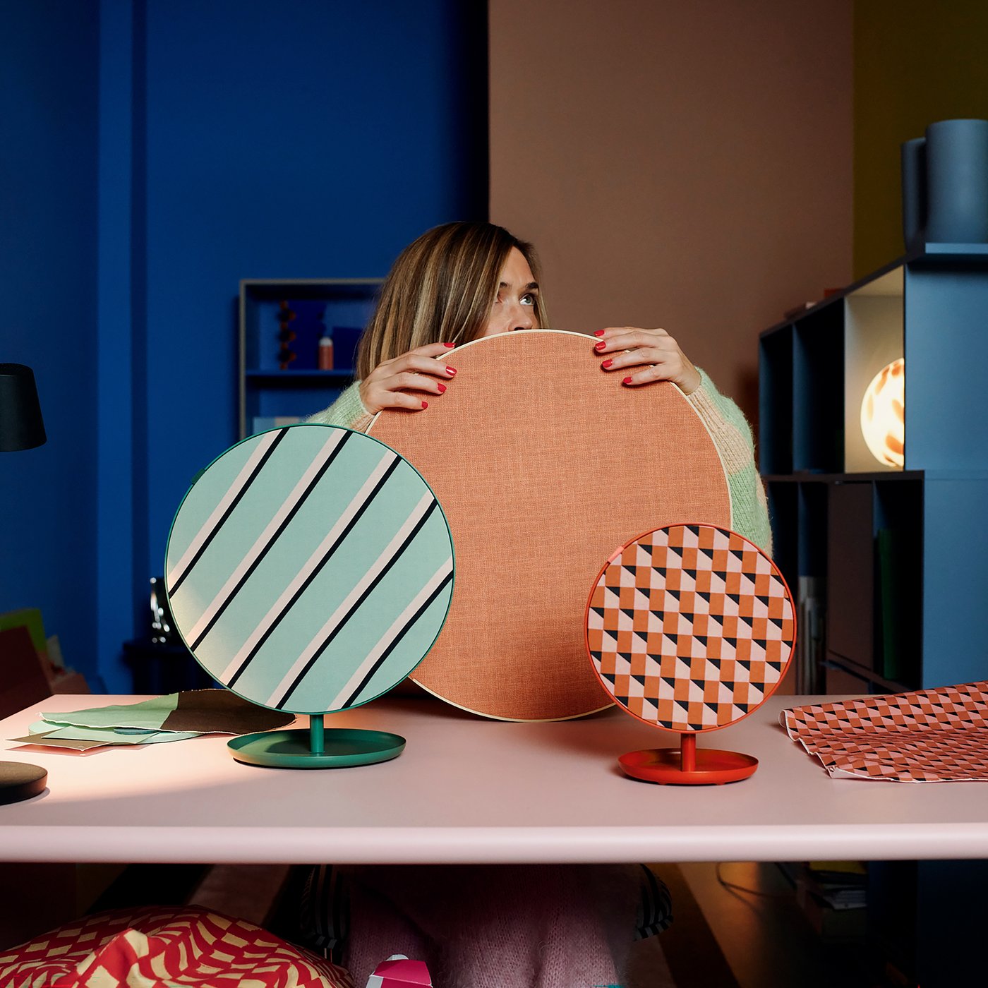 Una persona tiene un grande oggetto rotondo arancione con un cerchio blu, mentre due oggetti rotondi più piccoli, uno a strisce verdi e l'altro con un motivo geometrico arancione, sono su una scrivania rosa. Sullo sfondo si intravedono scaffali colorati e una lampada da tavolo nera.