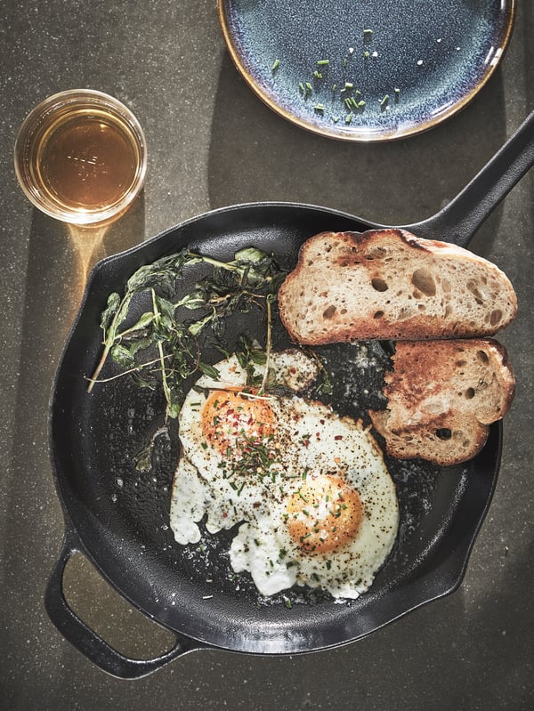 A VARDAGEN frying pan made of cast iron is placed on a dark surface and it has two fried eggs and two pieces of toast in it.