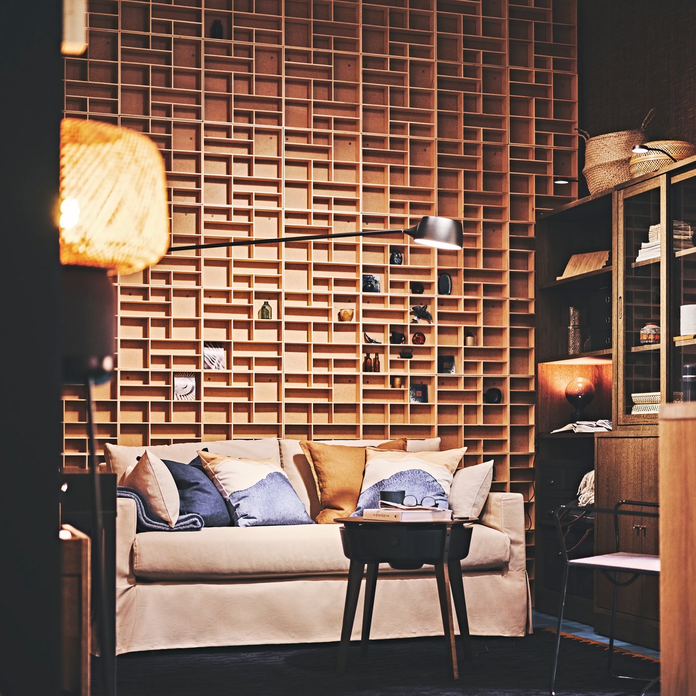 Vardagsrum med beige soffa, blå och beige kuddar, träbord, trävägg med mönster och hylla med böcker och korgar.