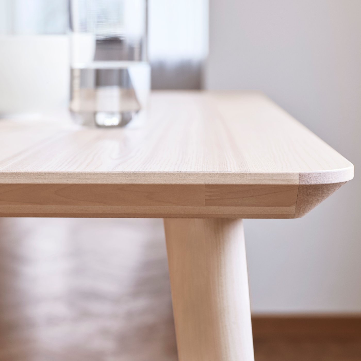 The smooth edge of a light-coloured LISABO coffee table highlighting the ash veneer material, with a glass of water on top.