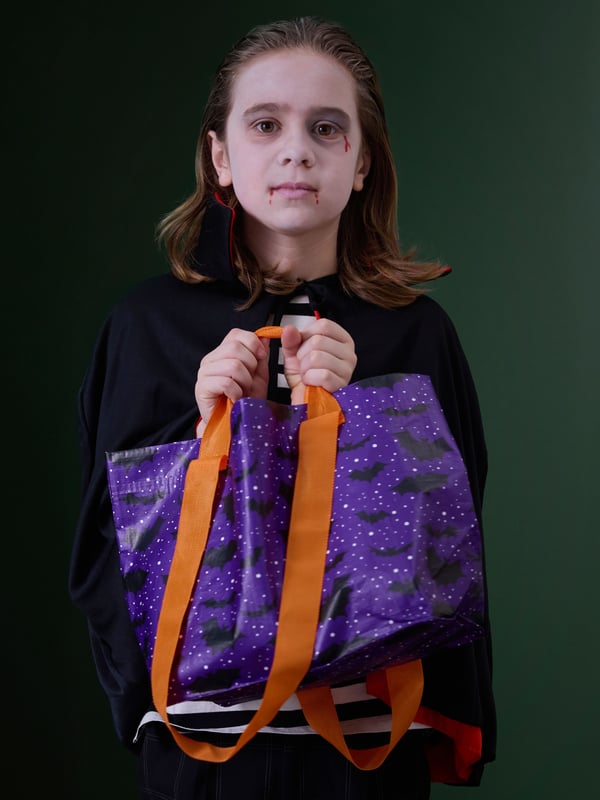 A kid dressed up in a black-and-white clown costume holds a HÖSTAGILLE bag with a bat pattern.