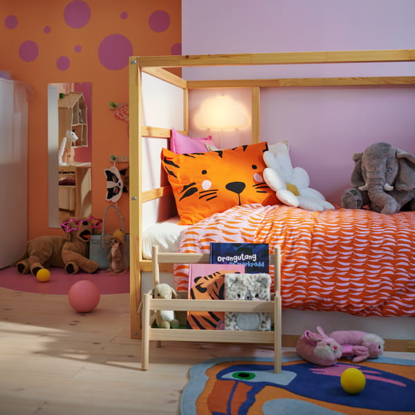 A KURA reversible bed in a colourful children’s bedroom with soft toy animals on it and a FLISAT book display in front.