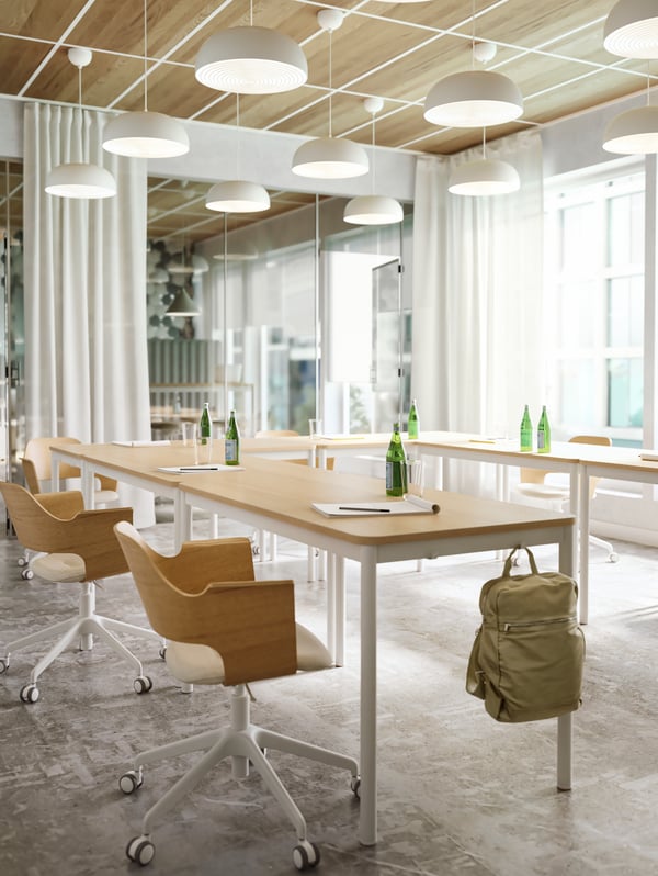 Chairs in light wood, white lamps and tables in light wood with hooks on the side where a green backpack is hanging.