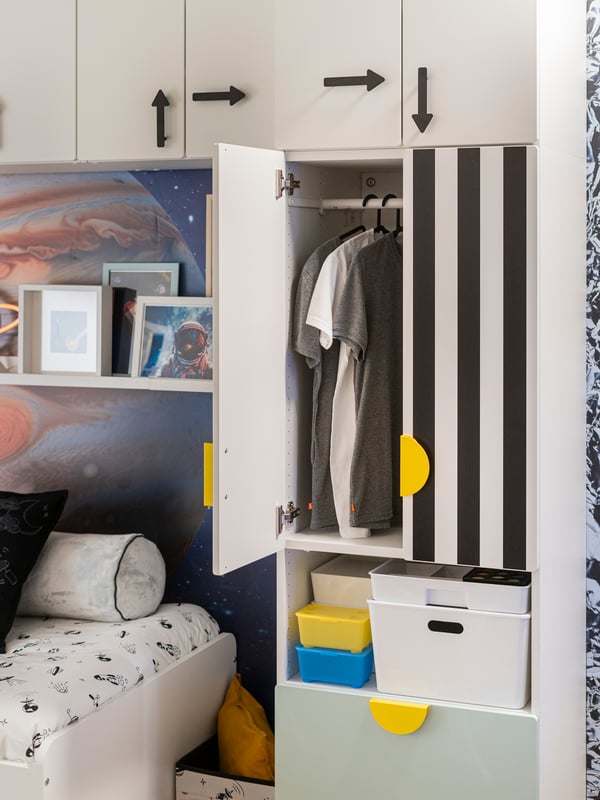 Un anta di un guardaroba SMÅSTAD con un anta aperta che svela delle magliette appese a un bastone SPRUTTIG. Nel ripiano inferiore diversi contenitori.