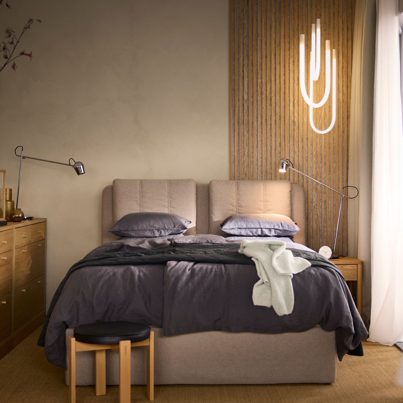 Modernes Schlafzimmer mit einem gepolsterten Doppelbett in Beige, grauer Bettwäsche und einer dekorativen Holztrennwand mit weißem grafischem Muster. Links steht eine Holzkommode, rechts ein kleiner Holzstuhl, und die Beleuchtung ist warm und gemütlich.