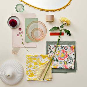 A folded yellow and white FJUNKLOCKA cushion cover placed among other items, flowers, and textiles in a stylish arrangement.