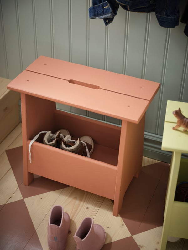 A close up of a PERJOHAN bench painted rust red shows a pair of kid shoes stored in the shelf underneath.