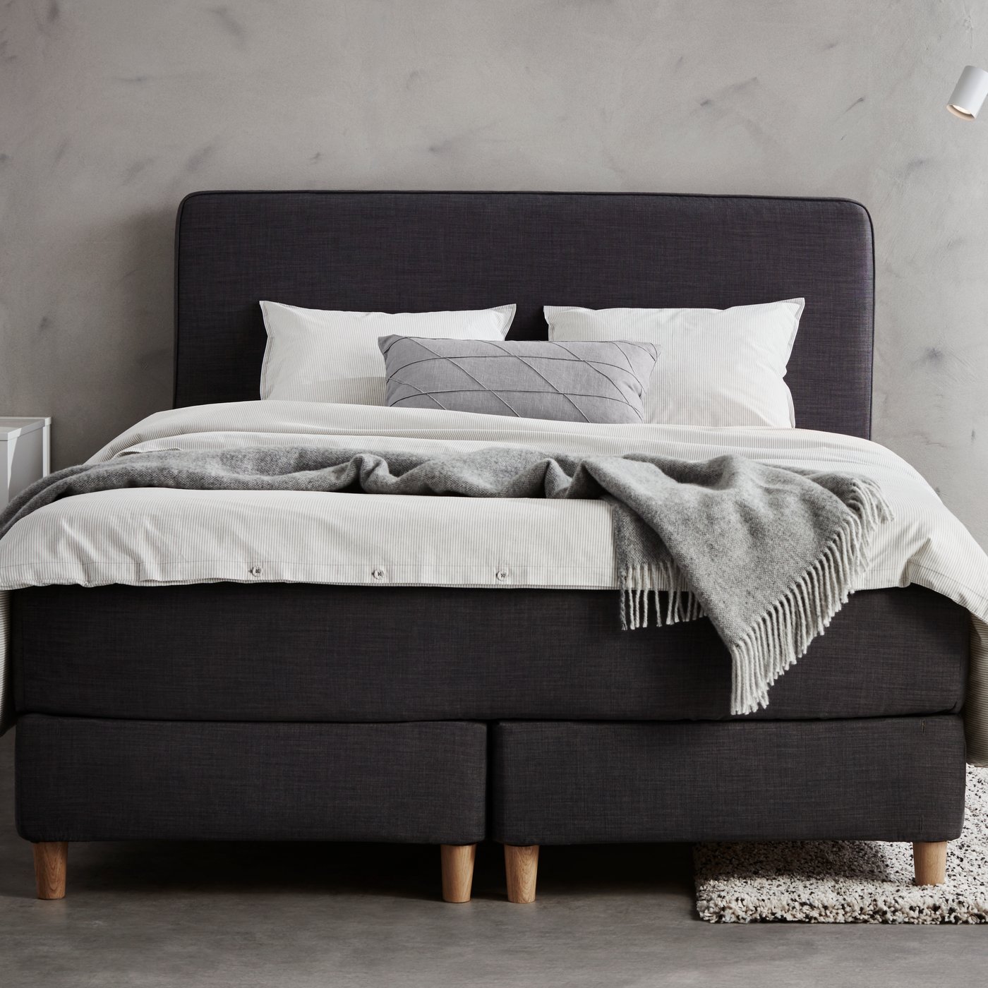 Ein schwarzes DUNVIK Bett steht an einer weißen Wand. Darauf liegen graue und weiße Pölster und eine graue Decke.