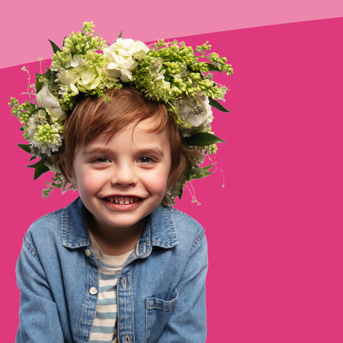 A smiling boy wearing a denim jacket, a striped T-shirt, and a large white and green flower crown, positioned in front of a two-tone pink background.