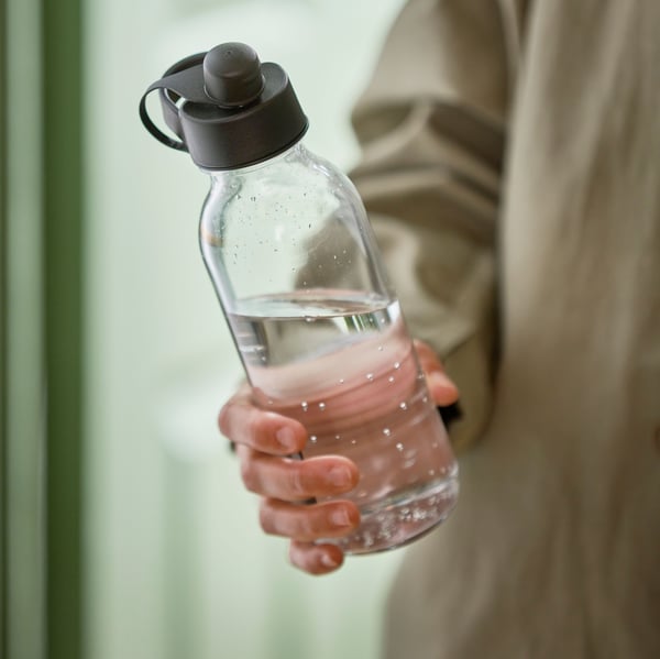 A person in a beige sweater is holding an IKEA 365+ water bottle with a dark grey lid which is half filled with water.