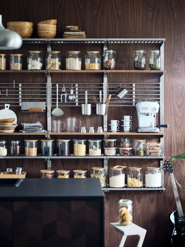Offene KUNGSFORS Küchenregale aus Edelstahl und Holz an dunkler Holzwand, ordentlich bestückt mit Vorratsgläsern, Tassen, Küchenutensilien und weißer Espressomaschine.