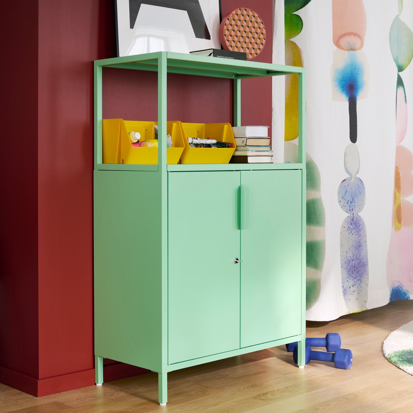 A bright room features a light-green TROTTEN cabinet with doors, holding supplies and books beside colourful wall art.
