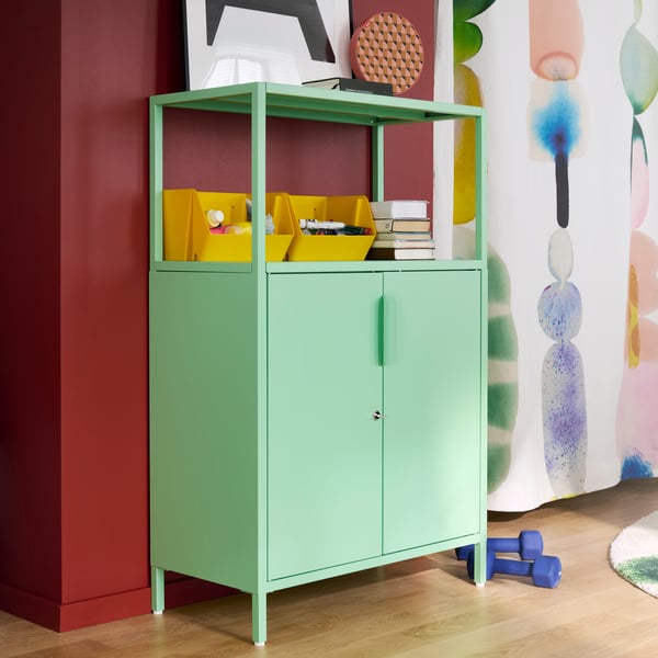 A bright room features a light green TROTTEN cabinet with doors, holding supplies and books beside colourful wall art.