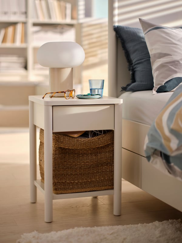 Uma mesa de cabeceira TONSTAD em branco sujo, com um candeeiro de mesa DEJSA em bege e branco opalino por cima, colocada ao lado da cama.
