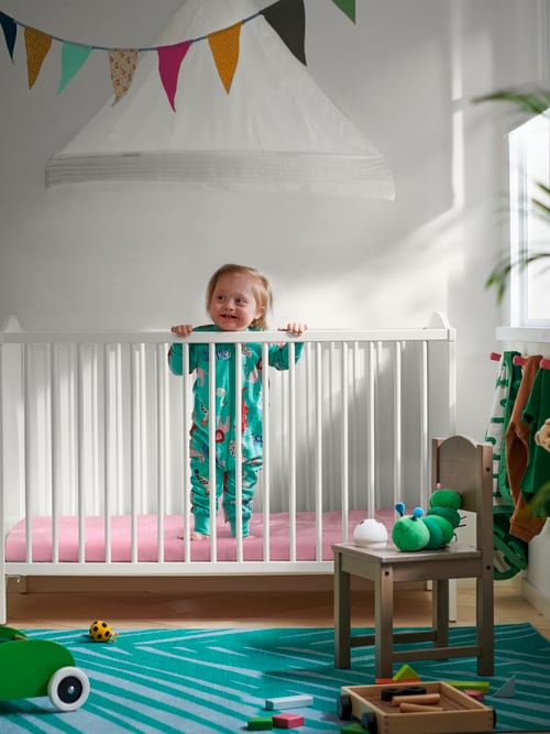 A child standing in a SMÅGÖRA cot with multicolour bunting on the wall above with a SUNDVIK children's chair in front.