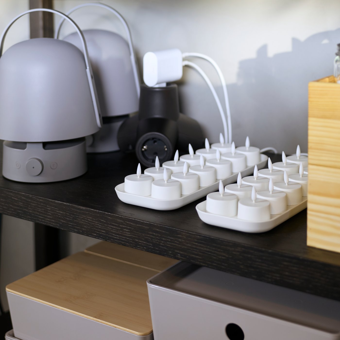 Bílá sada LED čajových svíček SKATGÅS stojí na polici spolu se dvěma lampami s reproduktorem a s nabíjecími zařízeními.