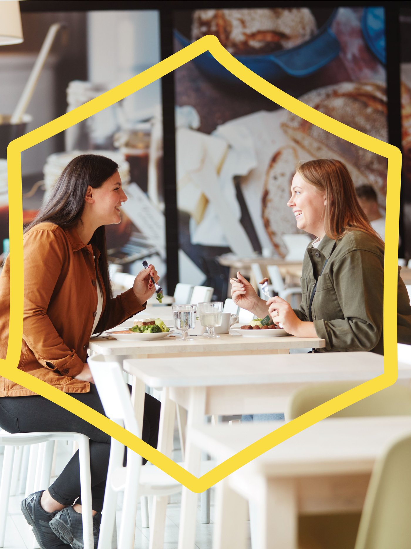 Zwei Frauen sitzen lachend in einem IKEA Restaurant und sind dabei etwas zu essen und zu trinken. 