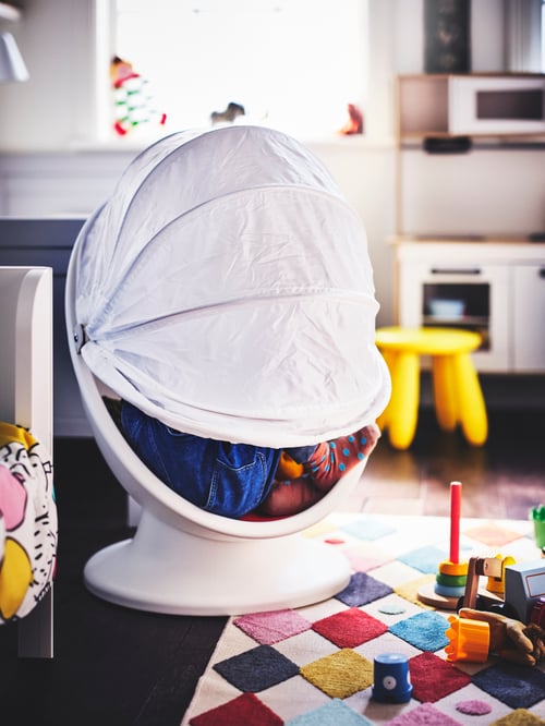 Children’s room with child hiding inside an egg-shaped white swivel chair with red cushion and foldable fabric hood.
