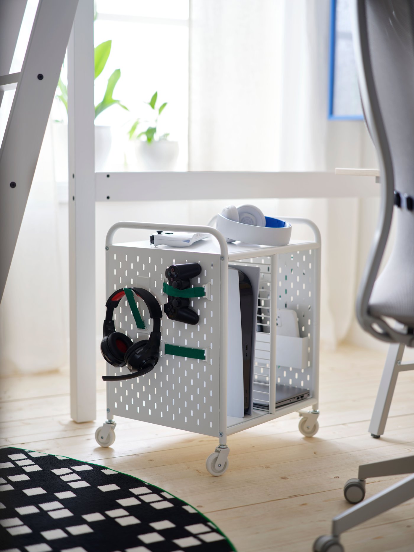 Una estantería BRÄNNBOLL blanca con ruedas sostiene auriculares y mandos en su lado de tablero de clavijas, junto a una alfombra y una silla de juegos.