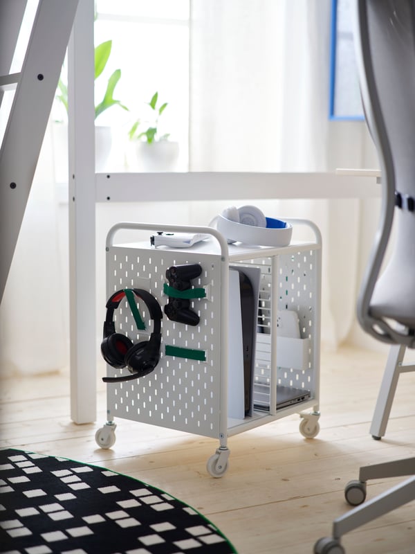 A white BRÄNNBOLL shelving unit on castors holds a headset and controllers on its pegboard side, by a rug and a gaming chair.