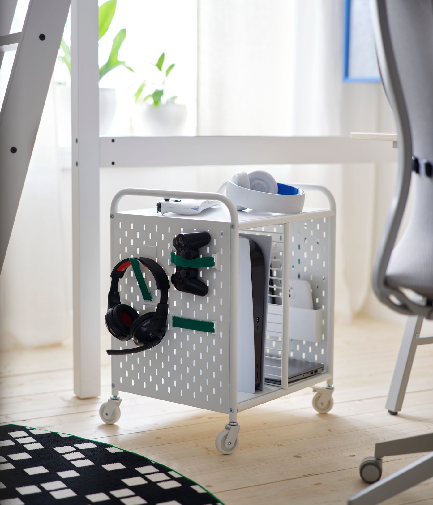 A white BRÄNNBOLL shelving unit on castors holds a headset and controllers on its pegboard side, by a rug and a gaming chair.