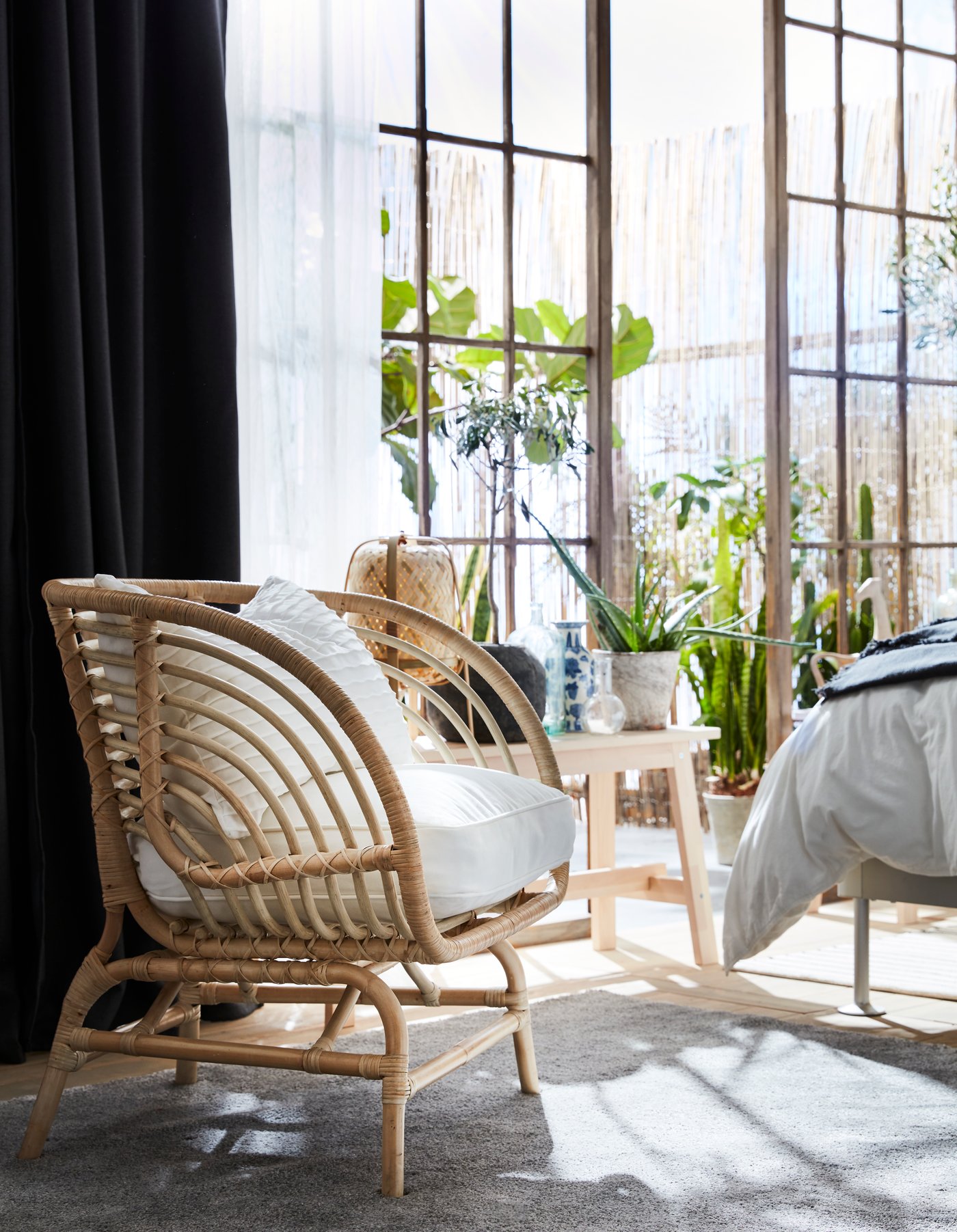 BUSKBO rottingfåtölj med vit dyna står vid ett ljust fönster med svarta gardiner, bredvid ett litet bord och gröna växter som skapar en luftig och naturlig känsla.