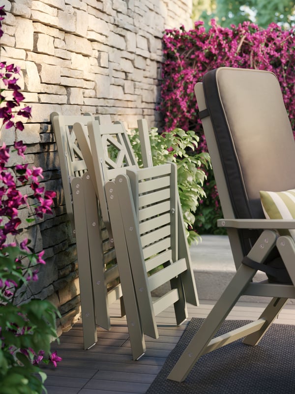 Two beige chairs folded up against a brick wall with purple vine and green plants and behind a reclining chair.