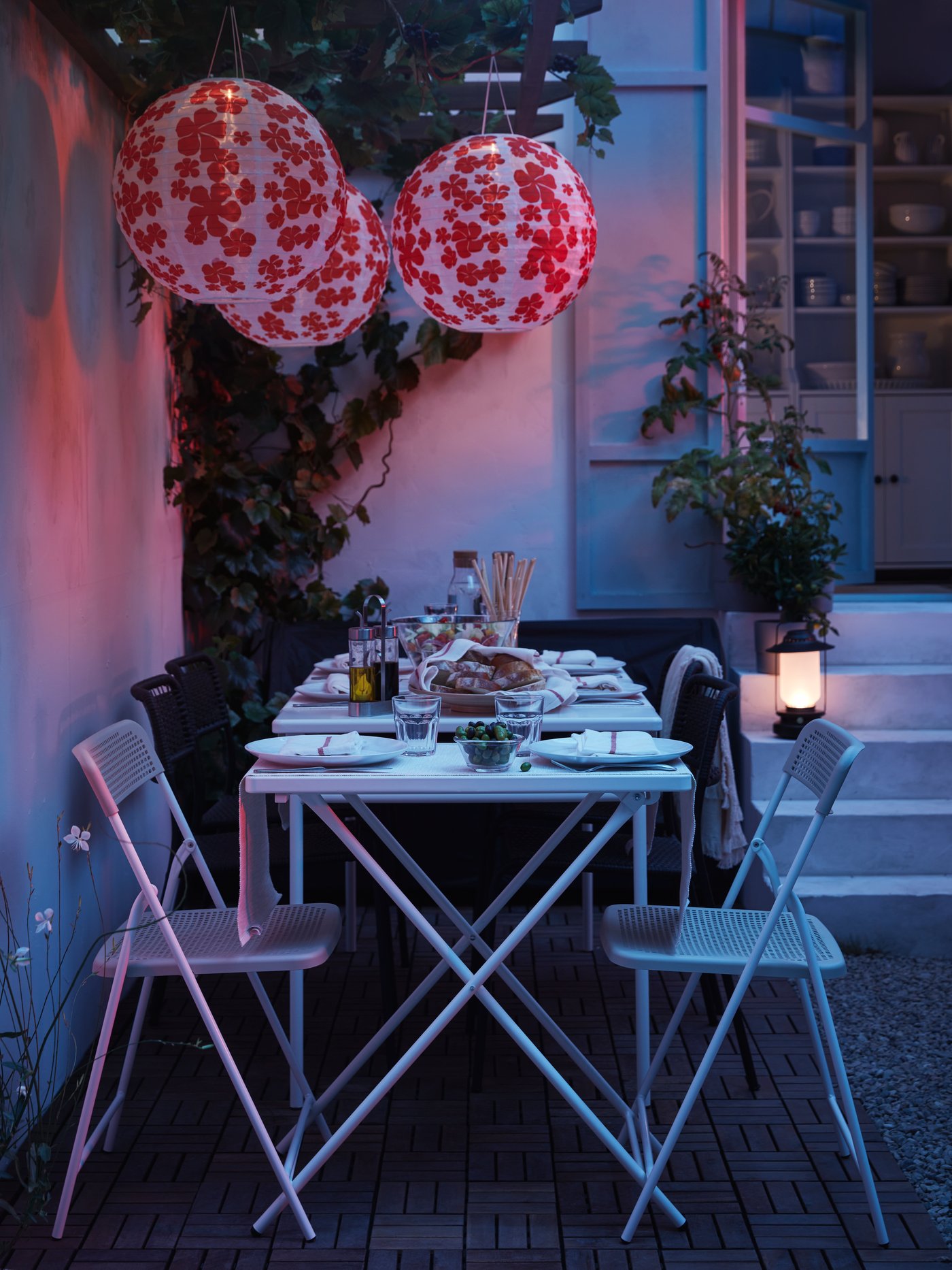 At night, a dinner party set up with two tables, four black chairs, two white chairs and three paper lanterns.