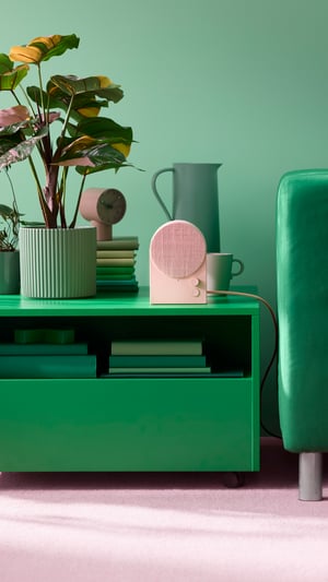 A dusty pink NATTBAD Bluetooth speaker on a green coffee table next to a green ribbed flowerpot containing a houseplant.