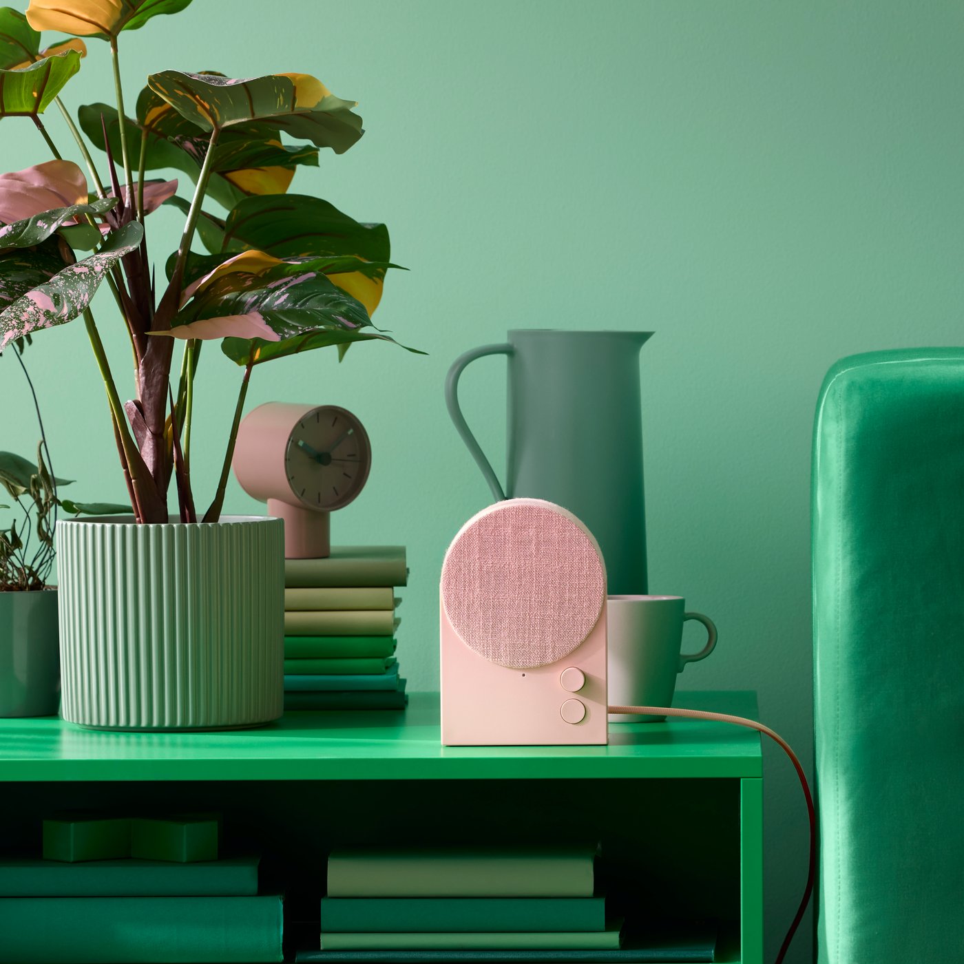A dusty pink NATTBAD Bluetooth speaker on a green coffee table next to a green ribbed flowerpot containing a houseplant.