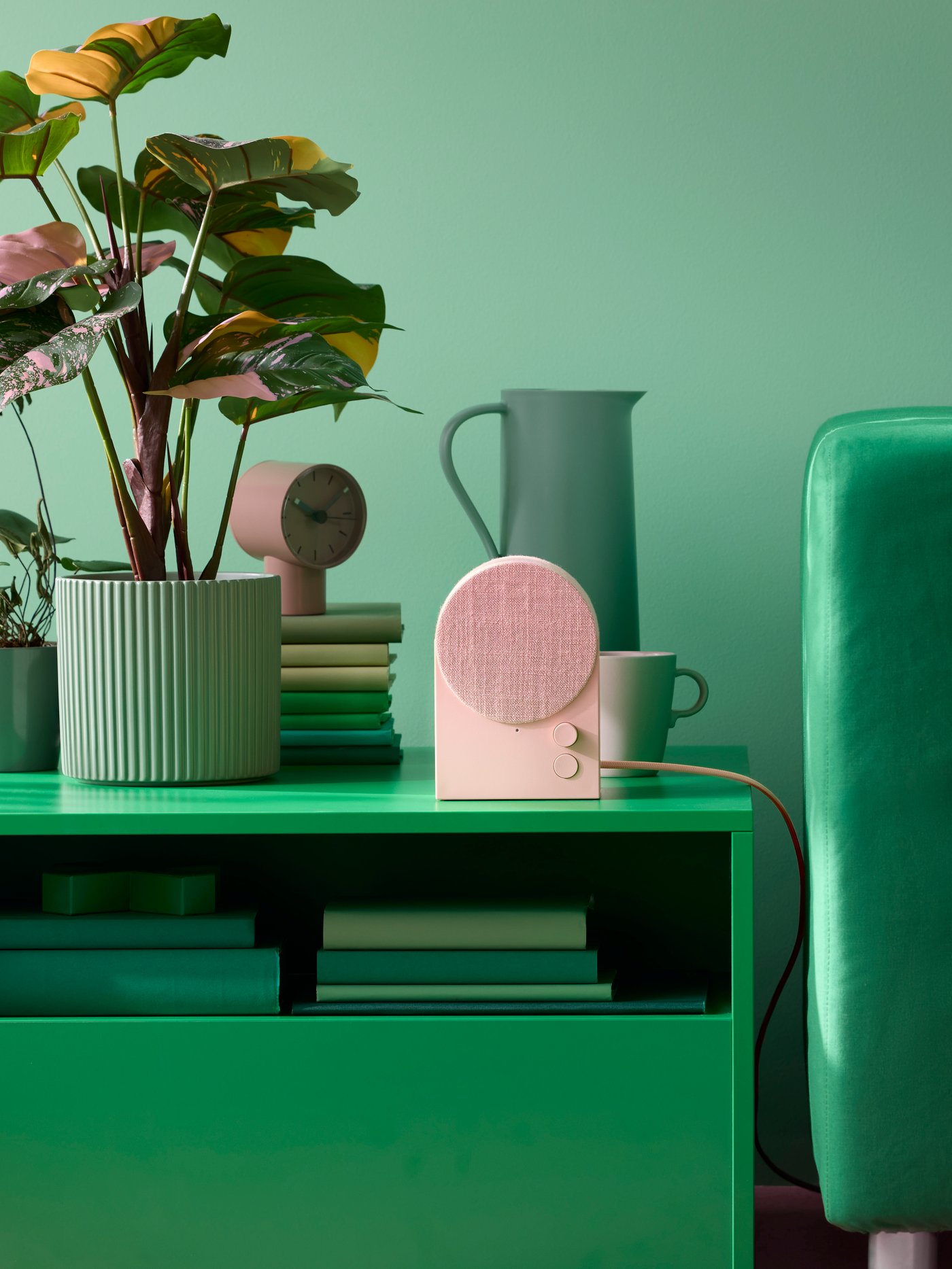 A dusty pink NATTBAD Bluetooth speaker on a green coffee table next to a green ribbed flowerpot containing a houseplant.