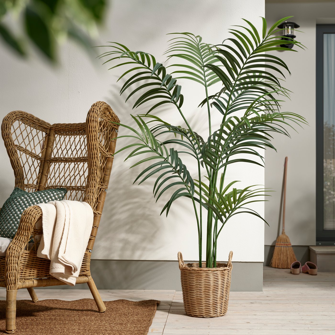 Uno spazio esterno con una pianta artficiale FEJKA da interno/esterno/Kentia forsteriana in vaso accanto ad una poltrona RISHOLMEN marrone.