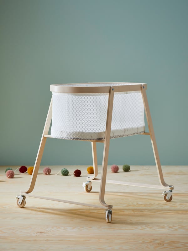 An ÄLSKVÄRD bassinet with sleeping pad in birch in a room with wooden floors and grey/green walls.