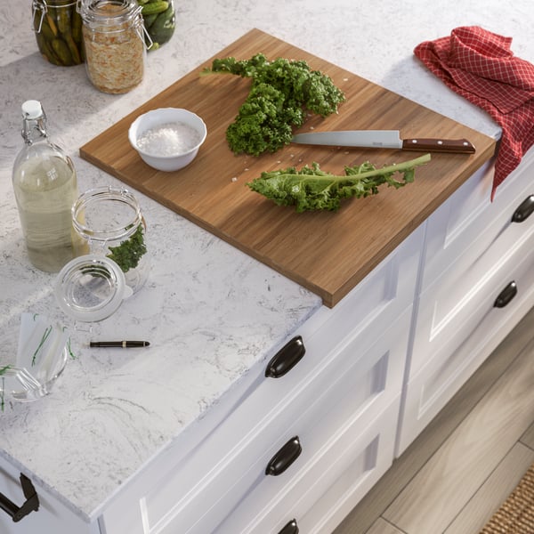 A cutting board with fresh kale and a knife on a quartz kitchen countertop, alongside a bottle and jars.