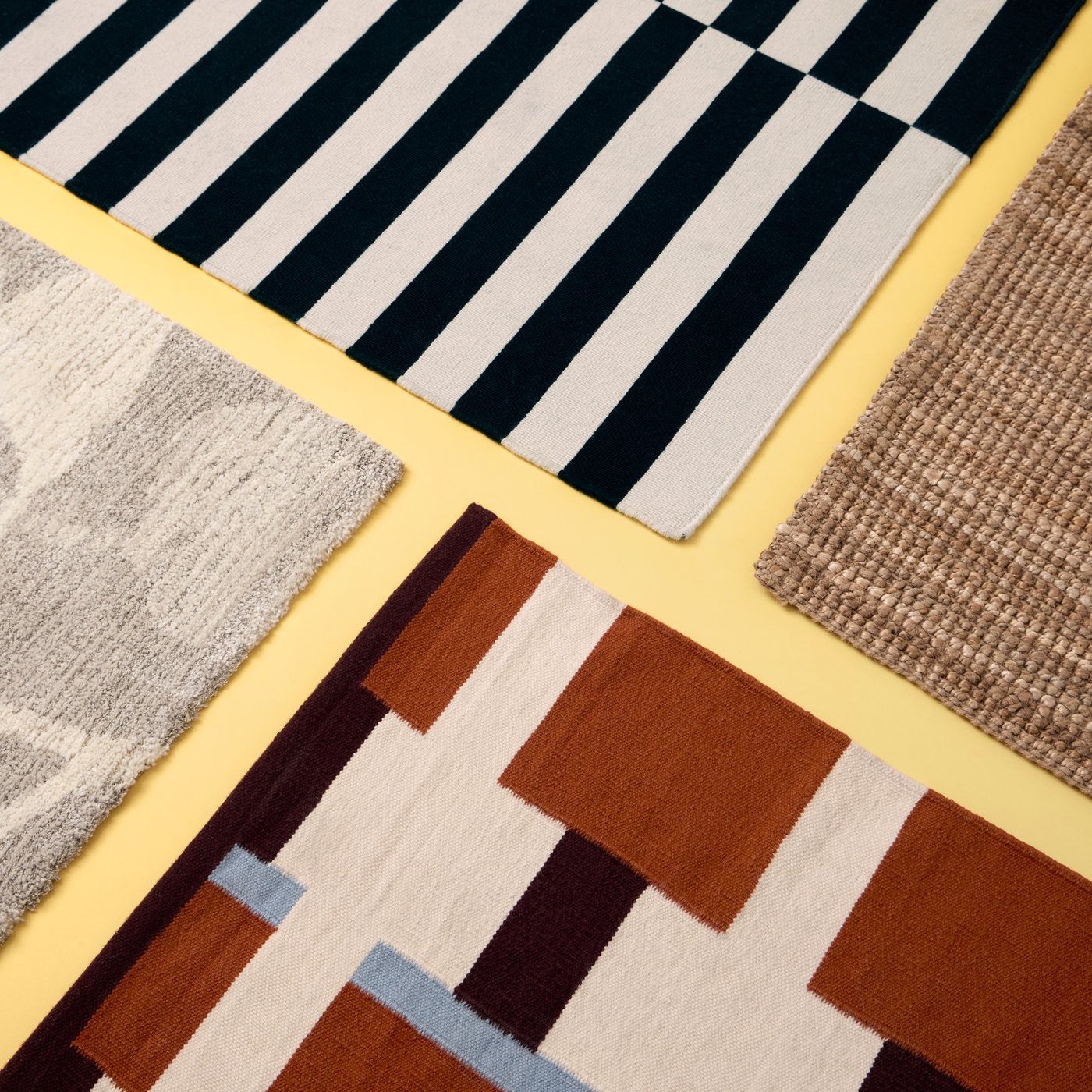 An arrangement of four rugs, one of them a white-brown-light-blue STOCKHOLM flatwoven rug, placed on a pastel-yellow surface.